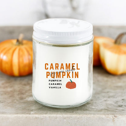 A white candle jar with 'CARAMEL PUMPKIN CARAMEL VANILLA' printed on the label, placed on a surface with small pumpkins in the background.