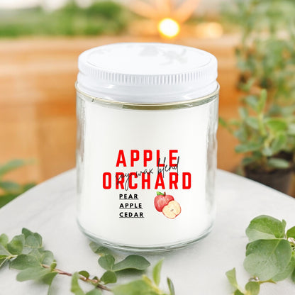 A white candle with red and green label that reads 'Apple Orchard', featuring images of an apple and a pear, set on a surface with green leaves around it.