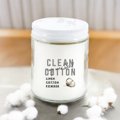 A white candle labeled 'Clean Cotton' with a depiction of a cotton plant, indicating the fragrance of linen, cotton, and powder.