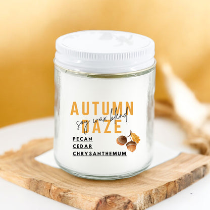 A white candle jar with a label that reads 'Autumn Daze' and fragrance notes 'Pecan, Cedar, Chrysanthemum'. The jar is placed on a wooden surface and has a white lid.