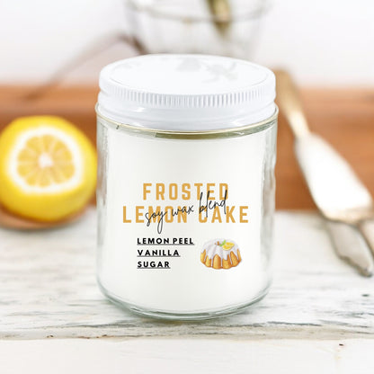 A white candle jar labeled 'FROSTED LEmon CAKE' with lemon peel vanilla sugar', depicting a lemon and vanilla cake image. The jar is on a wooden surface with a lemon and a silver candle snuffer in the background.