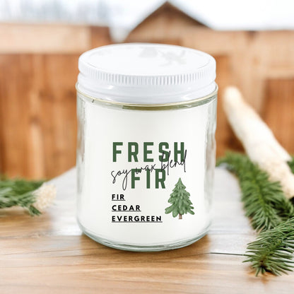 A white candle jar with a label reading 'FRESH FIR soy wax blend', featuring an illustration of a tree. The jar is on a wooden surface with greenery in the background.