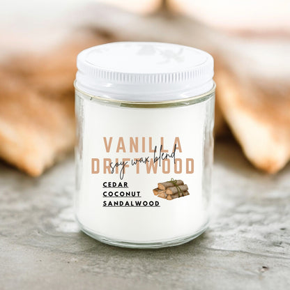 A white candle with the label 'Vanilla Driftwood' and fragrance listed as Cedar, Coconut, Sandalwood, placed on a surface with a warm, wooden background.