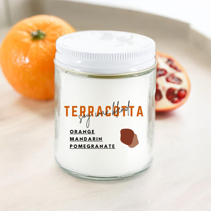 A white candle with the label 'Terracotta Orange Mandarin Pomegranate' on a kitchen counter with an orange and a pomegranate in the background.