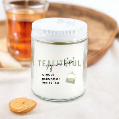A white candle jar with a label reading 'Tealiteful Ginger Bergamot White Tea', alongside a wooden lid and a lit candle in the background.
