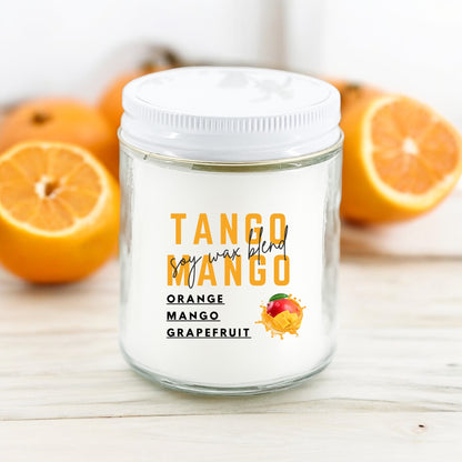 A white jar candle labeled 'Tango Mango' with orange and mango depicted in the label, placed on a wooden surface with halved oranges in the background.