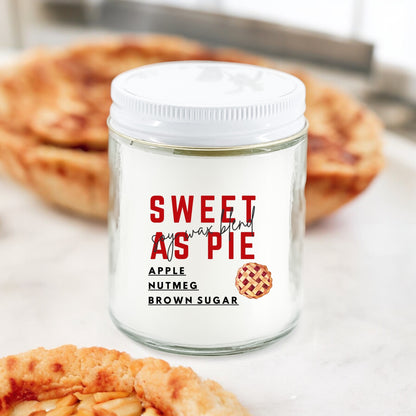 A white jar candle with the text 'SWEET AS PIE' and 'Apple Nutmeg Brown Sugar' on the label, placed on a surface with pie in the background.