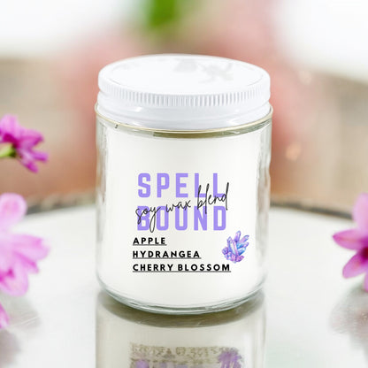 A white jar candle with the label 'Spell Bound' and scents listed as Apple, Hydrangea, Cherry Blossom. The candle is placed on a reflective surface with pink flowers around it.