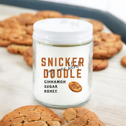 A white jar candle with the label 'Snickerdoodle' written on it, surrounded by cookies, with a wooden background.