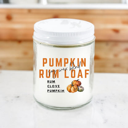 A white candle with a label reading 'PUMPKIN RUM LOAF' with images of a pumpkin and spices, placed on a kitchen countertop.