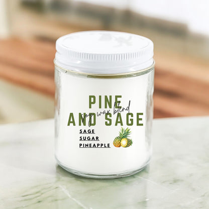 A white candle with the label 'Pine and Sage' and an illustration of a pineapple on it, placed on a marble surface.