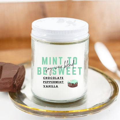 A white candle with green and brown text labeled 'Mint To Be Sweet Chocolate Peppermint Vanilla', placed on a glass dish with a piece of chocolate in the background.