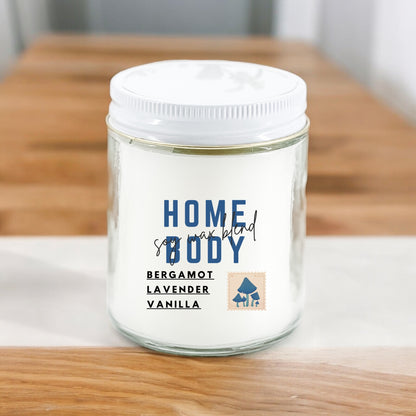 A white candle with the label 'Homebody' written on it, along with the scents 'Bergamot', 'Lavender', and 'Vanilla' listed below. The candle is placed on a wooden surface.