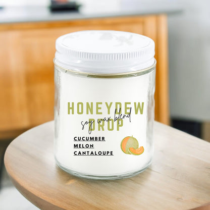A white candle with the label 'HONEYDEW DROP' and scents listed as Cucumber, Melon, and Cantaloupe. The candle is placed on a wooden surface.