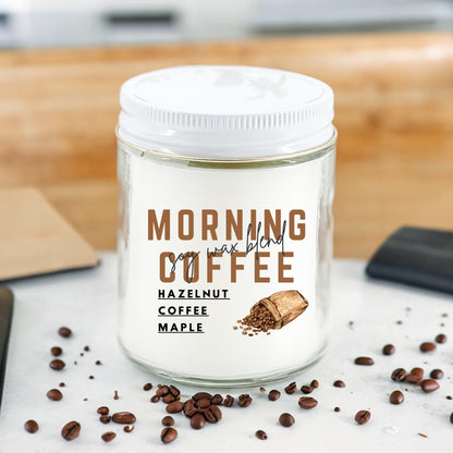 A white jar candle labeled 'Morning Coffee' with hazelnut and maple scents, surrounded by scattered coffee beans on a counter.