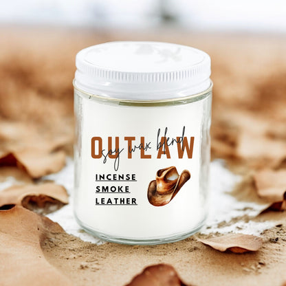A white candle with a label reading 'OUTLAW incense smoke leather' set on a bed of leaves, with a background of a wooden surface.