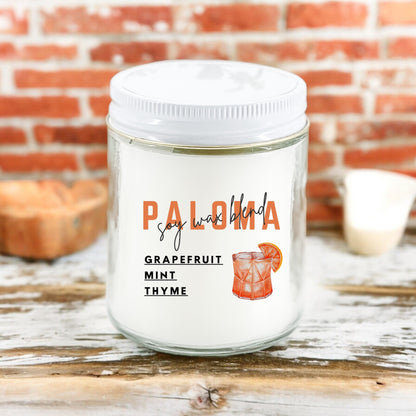 A white candle with the word 'PALOMA' printed in bold letters, and below it, 'Grapefruit Mint Thyme' written in smaller font. The candle is placed on a wooden surface with a brick wall in the background.