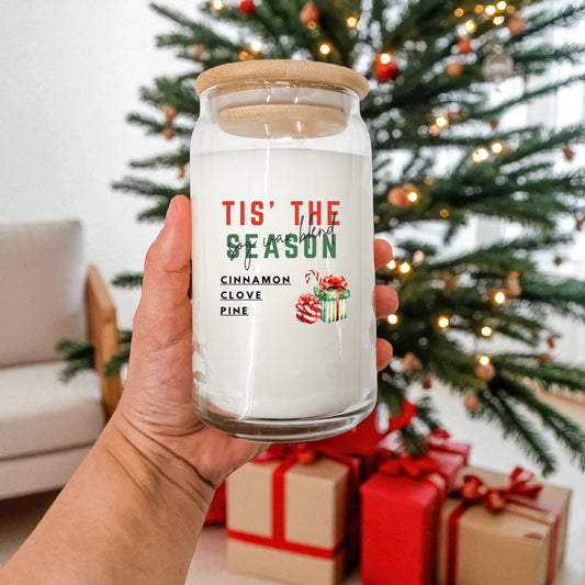 Hand holding a glass candle jar with Christmas-themed text and graphics in front of a decorated Christmas tree and presents.
