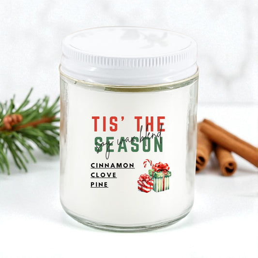 Glass candle jar with 'Tis' the Season label, cinnamon sticks, and pine branch on a white background